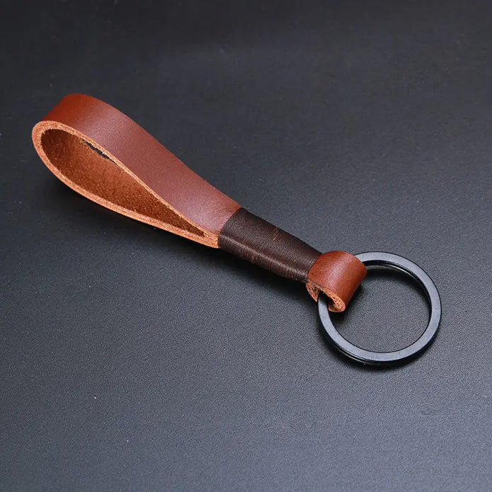 A brown, premium top-layer cowhide keychain featuring a robust, minimalist rope design. The keychain has a black metal ring and is placed on a neutral, dark gray background, showcasing the rich texture and color of the leather.