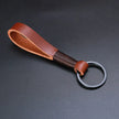 A brown, premium top-layer cowhide keychain featuring a robust, minimalist rope design. The keychain has a black metal ring and is placed on a neutral, dark gray background, showcasing the rich texture and color of the leather.