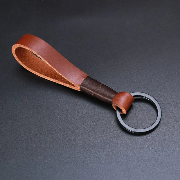 A brown, premium top-layer cowhide keychain featuring a robust, minimalist rope design. The keychain has a black metal ring and is placed on a neutral, dark gray background, showcasing the rich texture and color of the leather.
